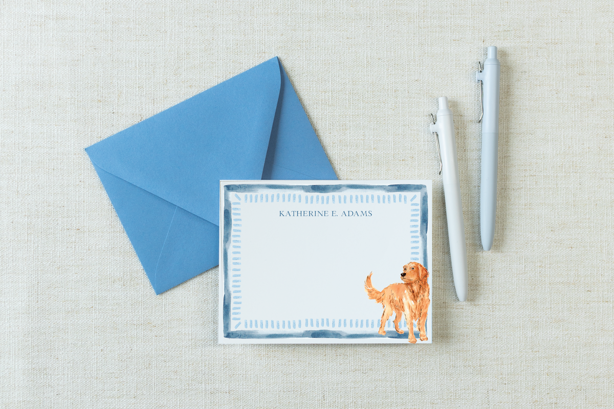 A personalized stationery card with a blue border and a watercolor golden retriever, shown with a matching blue envelope, a white pen, and a silver pen on a beige linen background.